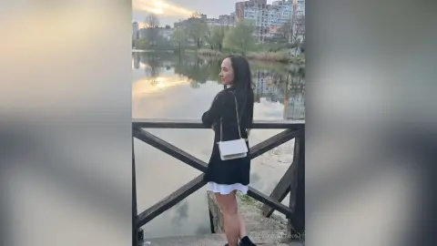 Young woman in a black dress and white purse standing by a lake at sunset.