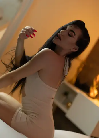 Sasha_Lorrenz poses in a tight, light-colored dress with her long dark hair flowing, showing off her shoulders and back in a warm-lit room.