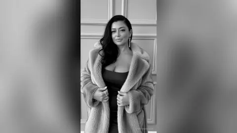 Elegant woman in a luxurious faux fur coat posing glamorously against a classic paneled wall background.