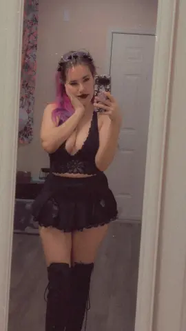 Young woman with pink hair in a black lace crop top and mini skirt taking a moody mirror selfie.