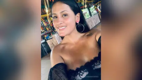 JulianaNavarro smiles while wearing a black lace top and large hoop earrings, showing her shoulders and long dark hair.