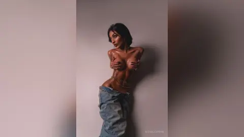 Glamorous woman in denim jeans and gold body chains posing against a neutral wall in moody studio lighting.