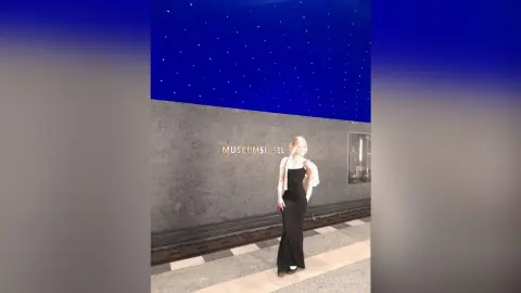 Elegant woman in a black evening gown and white blazer standing before the Museumsinsel building at night.