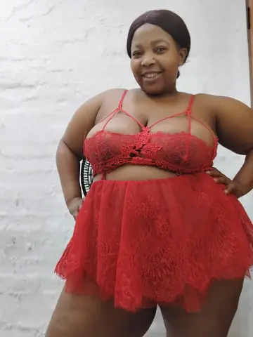 SexyChubbyxx29 smiles in a red lace lingerie set and short skirt while posing against a white brick wall.