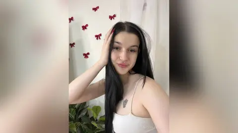 Young woman with long dark hair in a white tank top posing in a bright room with red bow decorations.