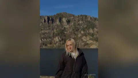 Blonde woman in a black puffer jacket standing before a scenic mountain lake under a clear blue sky.