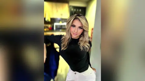 Smiling blonde woman in a black long-sleeve top and white pants posing in a modern kitchen setting.