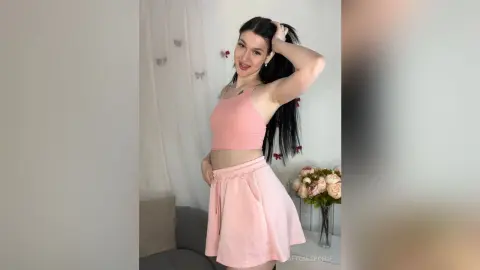 Smiling young woman in a pink ribbed crop top and matching skirt posing in a bright, airy room.