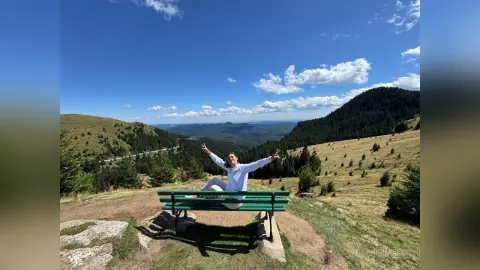 Cheerful woman in white outfit sitting on a park bench overlooking a scenic mountain landscape under a blue sky.