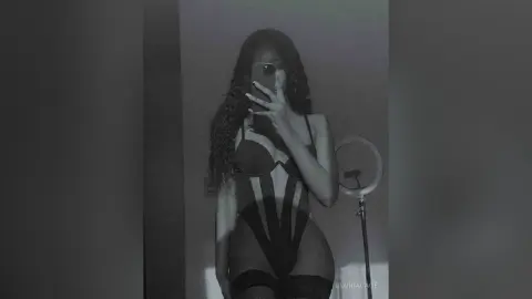 Curvy woman in a black cutout bodysuit posing for a mirror selfie in a moody, dimly lit room.