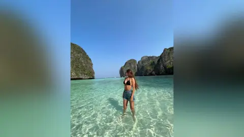 MilanaVolkova stands in clear shallow ocean water wearing a black bikini top and denim shorts, with her long brown hair down against a backdrop of tropical cliffs and blue sky.