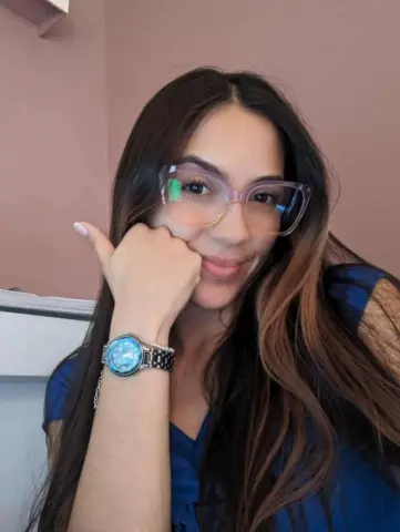 Smiling young woman with long brown hair and glasses wearing a blue shirt in a cozy indoor setting.