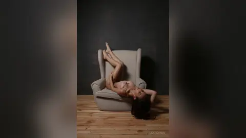 LeahEvans lies on her back in a grey armchair with her legs raised in the air against a dark background.