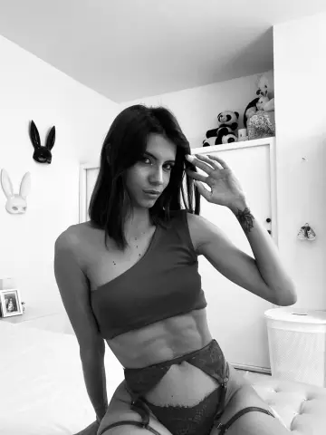 AprilJakovish poses in a red one-shoulder crop top and lace undies with garters, showing her midriff and dark hair in a bright room.