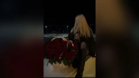 Blonde woman in a black outfit holding a large basket of red roses at night in a moody outdoor setting.