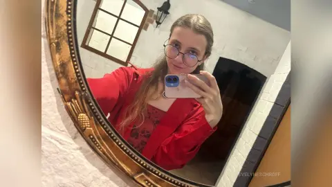 Young woman with glasses in a red shirt taking a selfie in a gold framed mirror indoors.