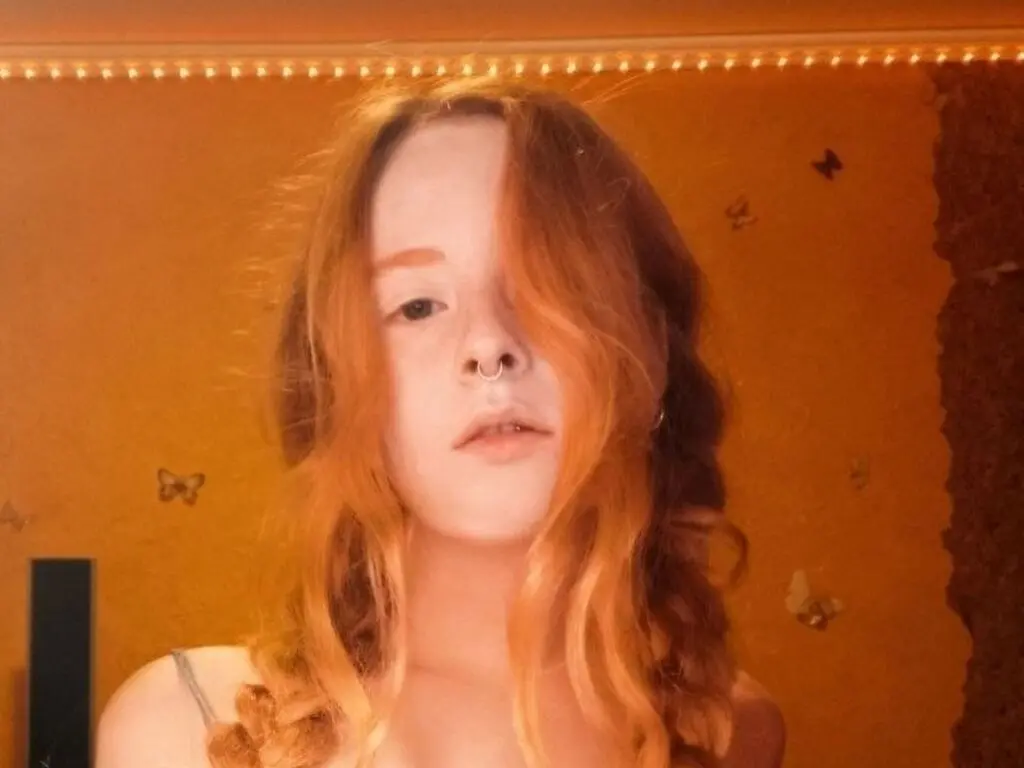 Young woman with wavy strawberry blonde hair and nose ring in warm, golden indoor lighting.