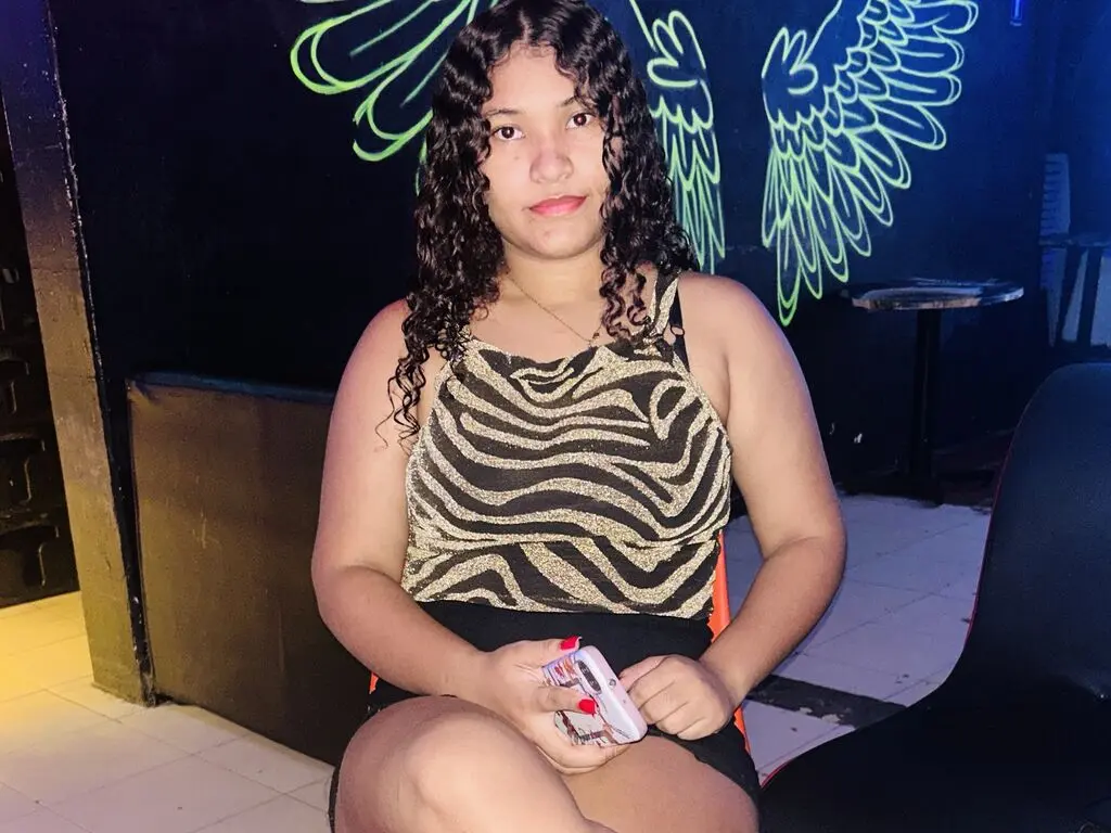 Young woman with curly hair in a gold zebra print top sitting in a dimly lit lounge with neon wing art.