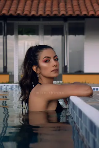 Young woman with long dark hair in a black swimsuit relaxing in a luxury swimming pool at sunset.