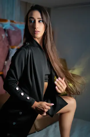 Elegant woman in a black blazer and crop top posing with flowing hair in a moody indoor setting.