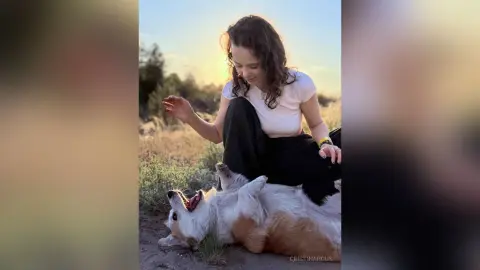 Smiling woman in a white t-shirt playing with a happy corgi in a sunlit grassy field at sunset.