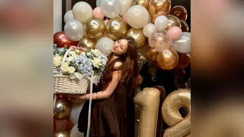 Smiling woman in a brown dress holding a flower basket amidst gold and white birthday balloons.