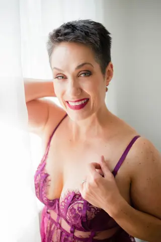 Smiling woman with short dark hair wearing a purple lace lingerie bodysuit by a bright window.
