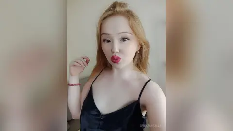 EpifaniaRalls pouts her red lips while wearing a black camisole, showing her long blonde hair and light skin against a plain background.