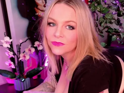 BlondeJJ — BlondeJJ, a blonde woman with blue eyes and pink lipstick, wearing a black top and a silver necklace, posing near white orchids and green leaves.