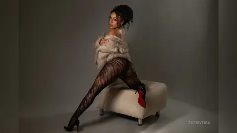 Glamorous woman in a fuzzy beige coat and patterned black tights posing on a white stool in a studio setting.