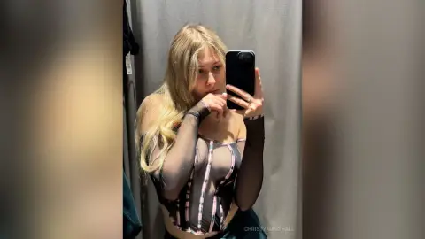 Blonde woman in a black sheer mesh bodysuit taking a mirror selfie in a fitting room.