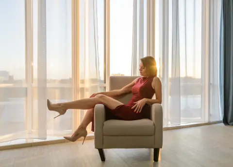 Elegant woman in a red cocktail dress sitting in a chair by a bright window in a sunlit modern room.