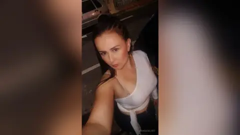 Young woman in a white tie-front top posing for a selfie on a city street at night.
