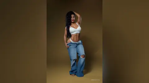 AnnyRoug poses in a white bra and low-rise ripped blue jeans, showing her toned stomach and midriff against a brown background.