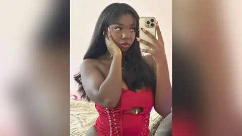 AmyJacobs poses in a red lace-up corset, holding a phone for a mirror selfie with her long dark hair down.