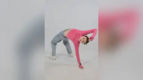 Cheerful woman in a pink long-sleeve top and light denim jeans stretching in a bright white studio.