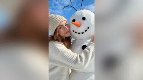 Smiling woman in a white knit sweater and beanie hugging a large snowman under a clear blue winter sky.