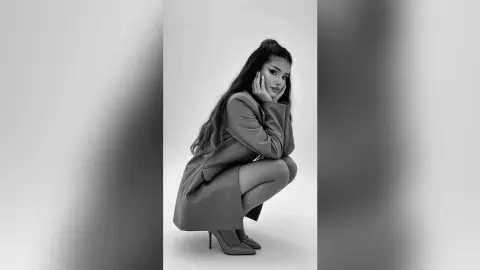 Melancholic woman in a long coat sitting on the floor against a minimalist white background.