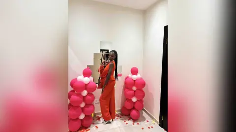 Stylish Black woman in a vibrant red jumpsuit posing next to pink and white balloon decor in a modern room.