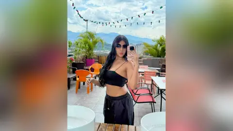 Young woman in a black crop top and shorts taking a selfie on a sunny rooftop terrace with mountain views.