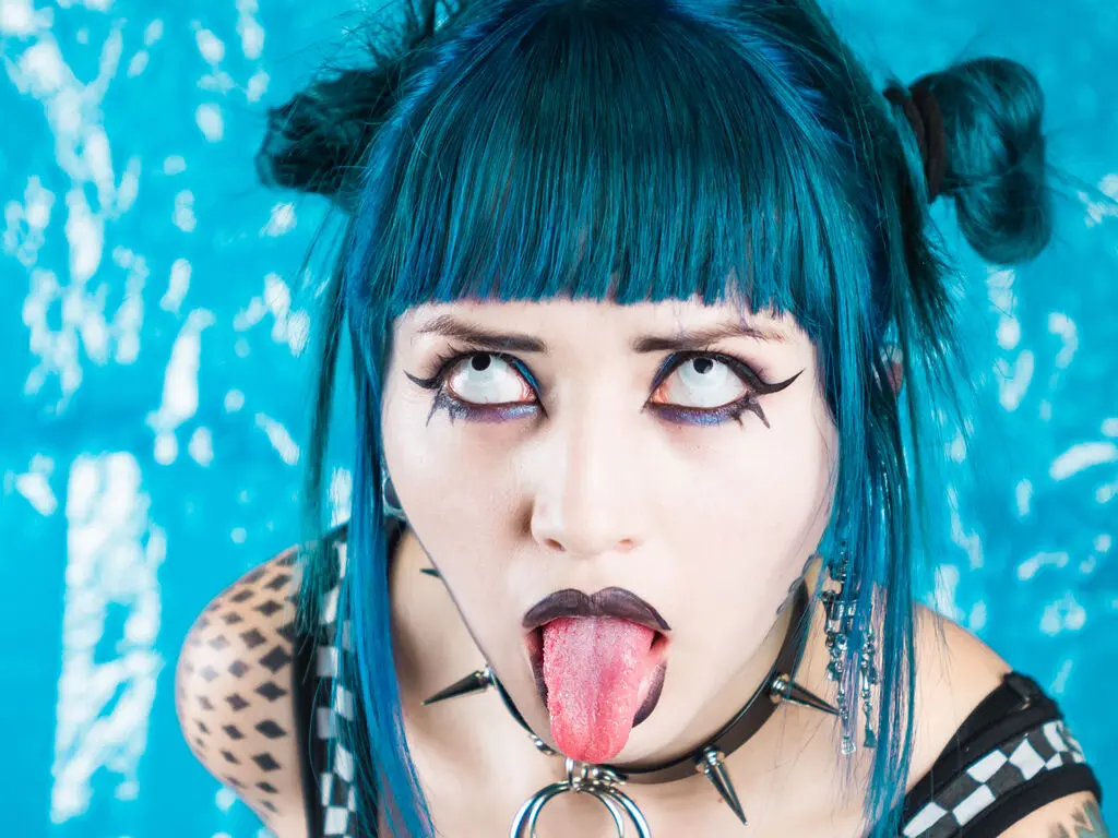 Woman with blue hair and gothic makeup wearing a black choker and studded outfit against a bright blue background.
