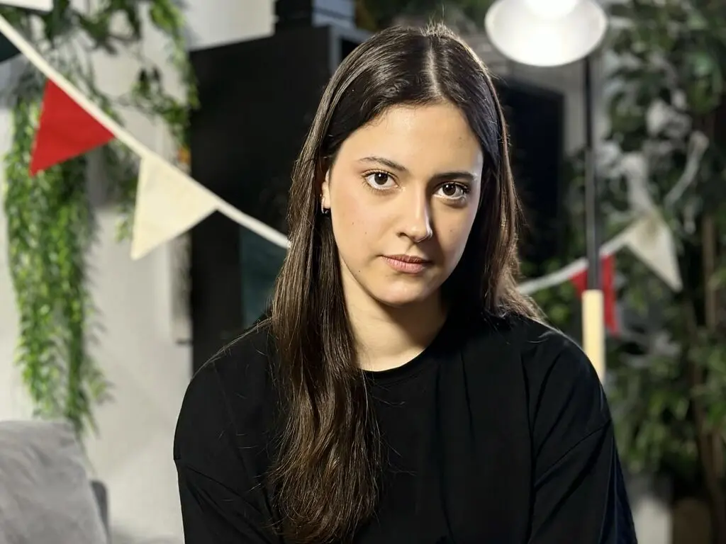 Young woman with long brown hair wearing a black t-shirt in a bright indoor setting with greenery.