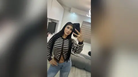 Young woman in a striped cardigan and blue jeans taking a selfie in a modern bedroom setting.