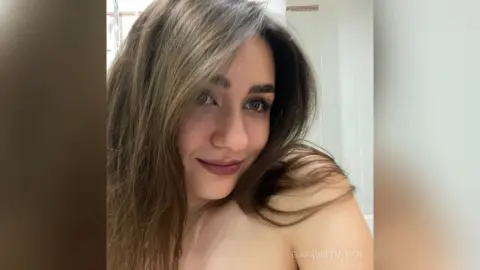 Smiling young woman with long brown hair posing in a bright, minimalist indoor setting.