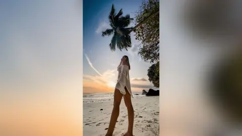 TheaSiena — Young woman in a white linen shirt standing on a tropical beach at sunset under palm trees.