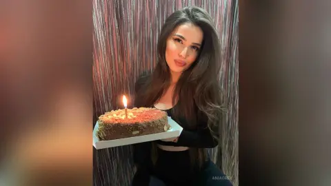 Young woman in a black top holding a chocolate birthday cake with a lit candle in a festive setting.