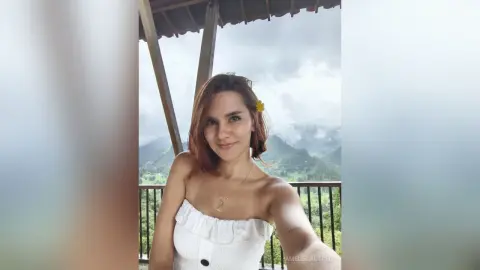 AmelieLautrec smiles while wearing a white strapless top and a yellow flower in her hair, standing on a balcony overlooking a lush, green mountain landscape.