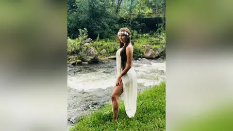 AndreaKalo poses by a rushing river wearing a long white dress and a white headband, showing off her long dark hair and bare legs.