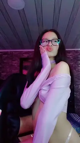 Young woman in glasses and a white off-the-shoulder top making a kissy face in a moody, dimly lit room.
