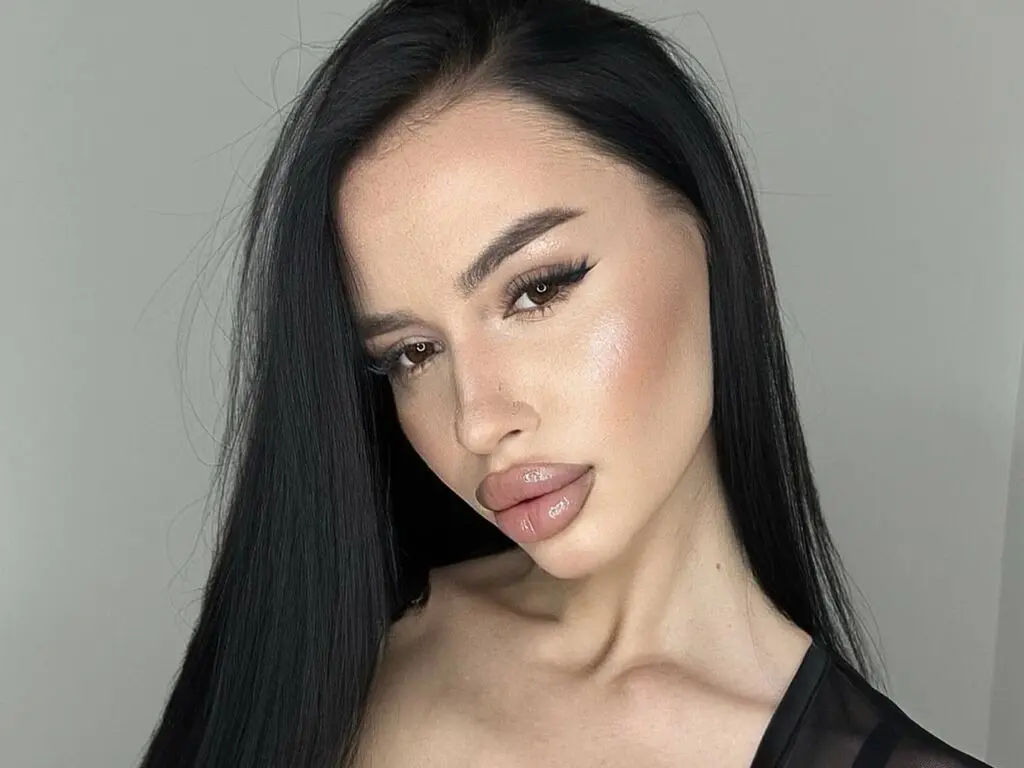 Young woman with long black hair and glowing makeup posing in a black outfit against a neutral background.